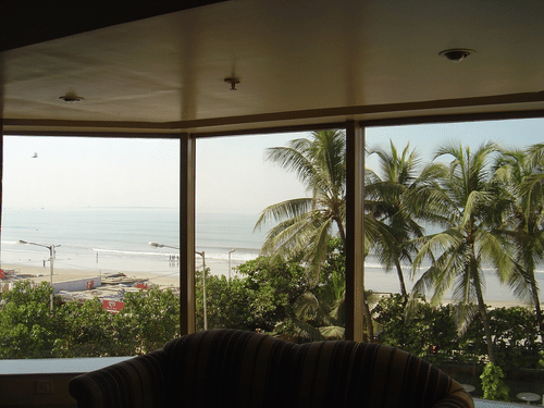 Scenic view of a beach and the sea through a big hotel room window, framed by lush palm trees.