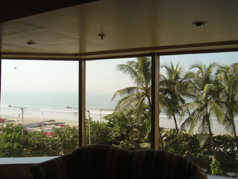 Scenic view of a beach and the sea through a big hotel room window, framed by lush palm trees.