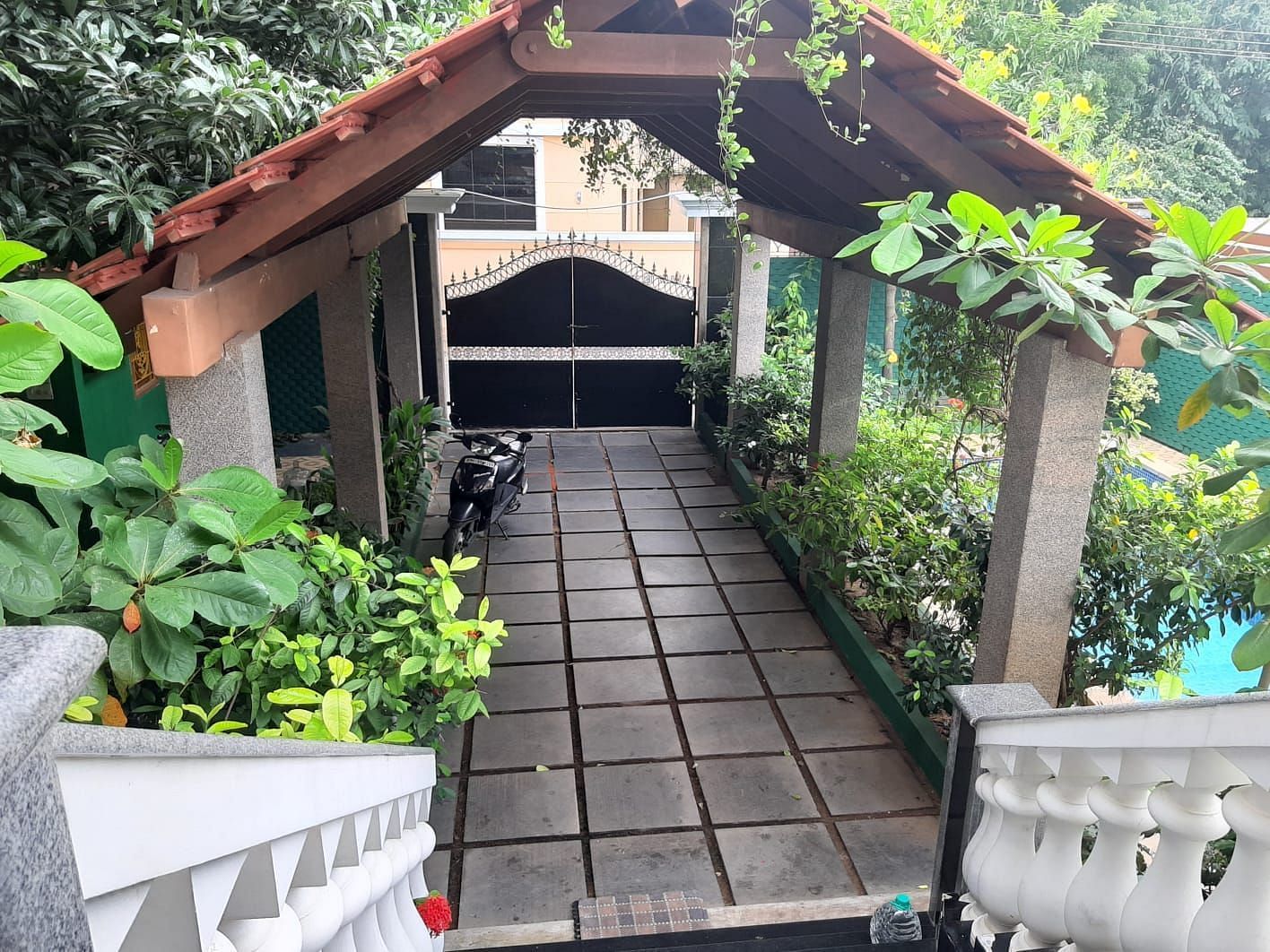 Covered outdoor walkway with a tiled path, surrounded by lush plants, leading to a gated entrance under a wooden roof structure.