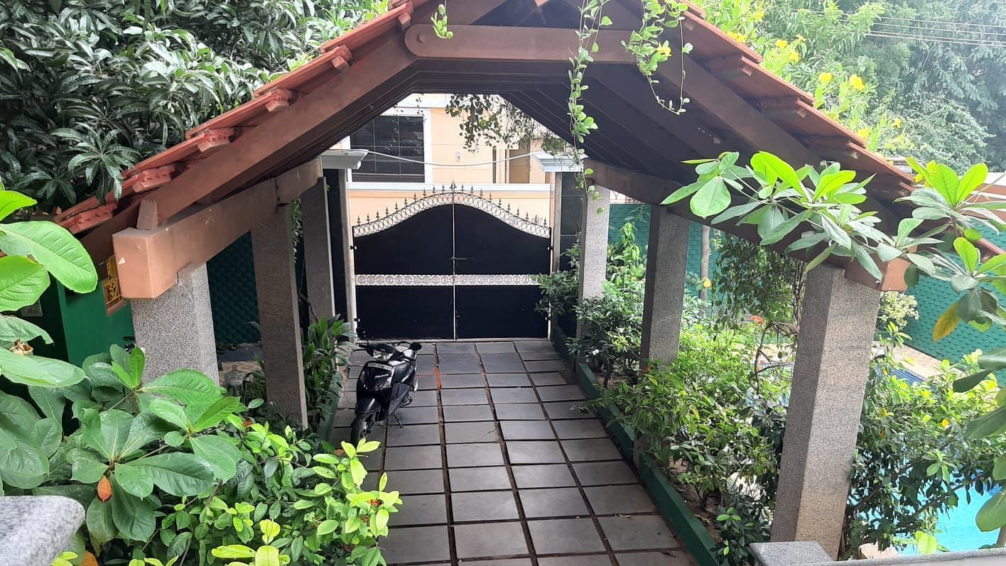 Covered outdoor walkway with a tiled path, surrounded by lush plants, leading to a gated entrance under a wooden roof structure.