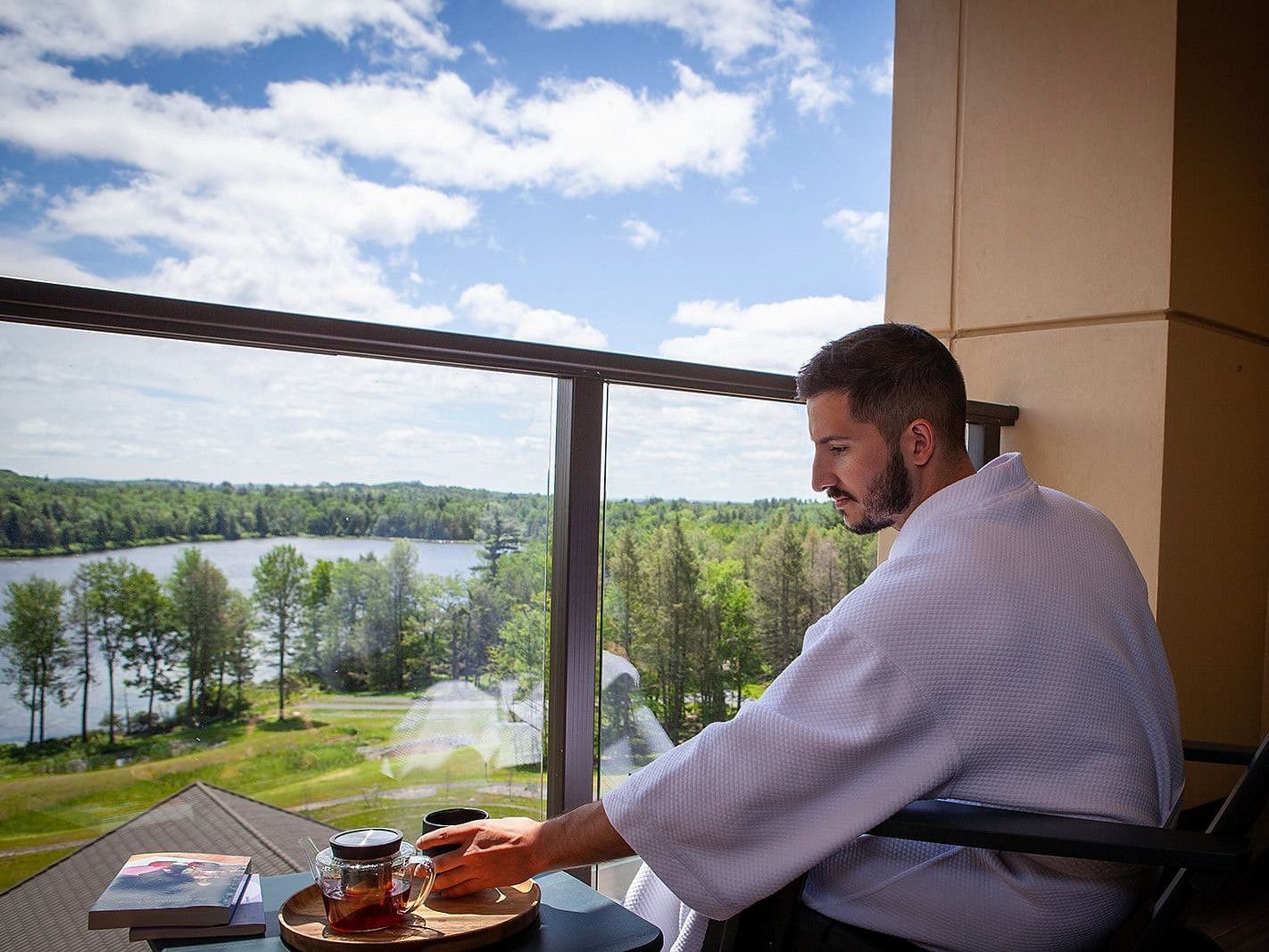 A man in bathrobe sitting on a chair in the balcony reaching out for a cup of beverage while enjoying the view outside at YO1 Longevity and Health Resorts in Catskills.