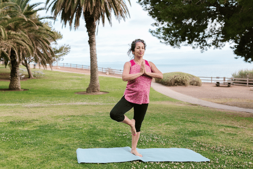 A woman is doing a tree pose on a blue yoga mat in a grassy park surrounded by lush trees.