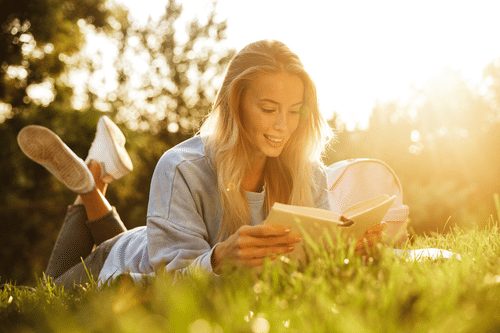 A smiling young woman with blonde hair lies on green grass, reading a book in the warm glow of the setting sun