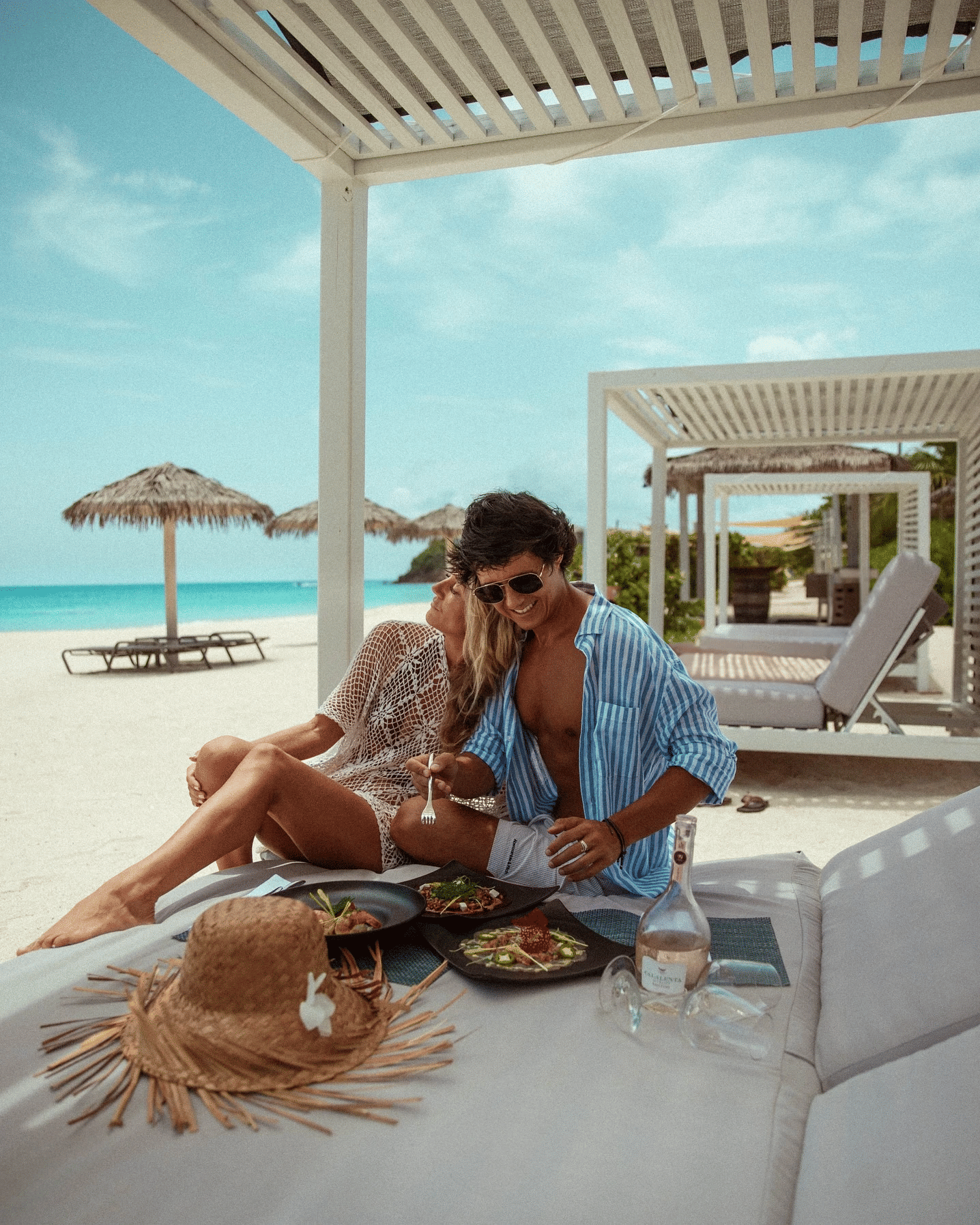 Two people enjoying a romantic beachfront meal under a white pergola with turquoise sea in the background.