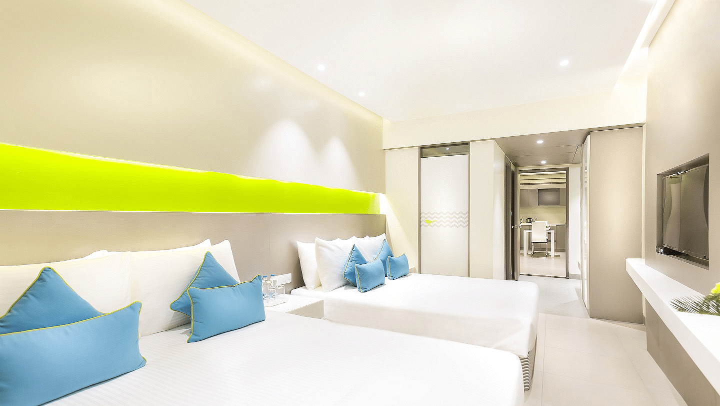 A spacious Suite features a large bed with a headboard stretching across the wall. Cushion chairs sit beside a wooden bedside table, and a wall-mounted television is positioned opposite the bed