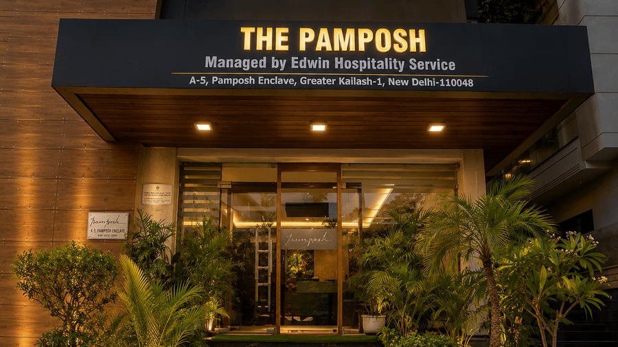The illuminated facade of The Pamposh, New Delhi, with plants and the sign board.