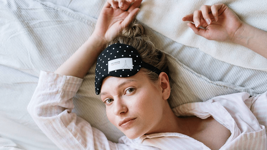 A woman wearing a sleep mask on her forehead resting on a bed with white linen