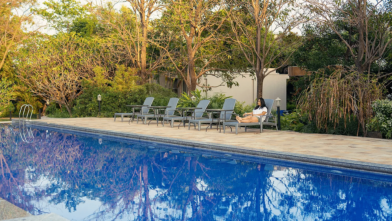 A calm swimming pool with several sun loungers kept besides the pool surrounded by greenery all around - Ivory Destinations