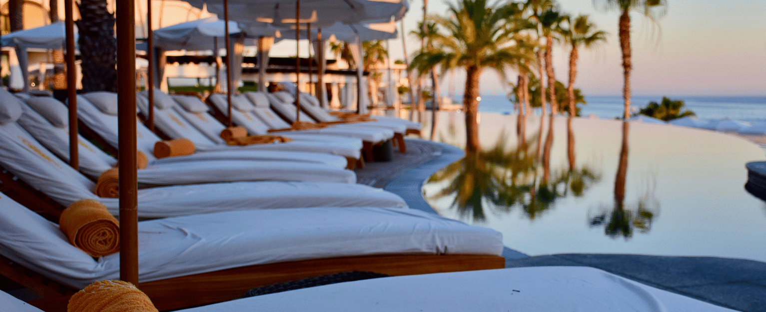 A serene waterfront view with luxurious poolside loungers overlooking a sunset-lit harbour, featuring yachts and palm trees.