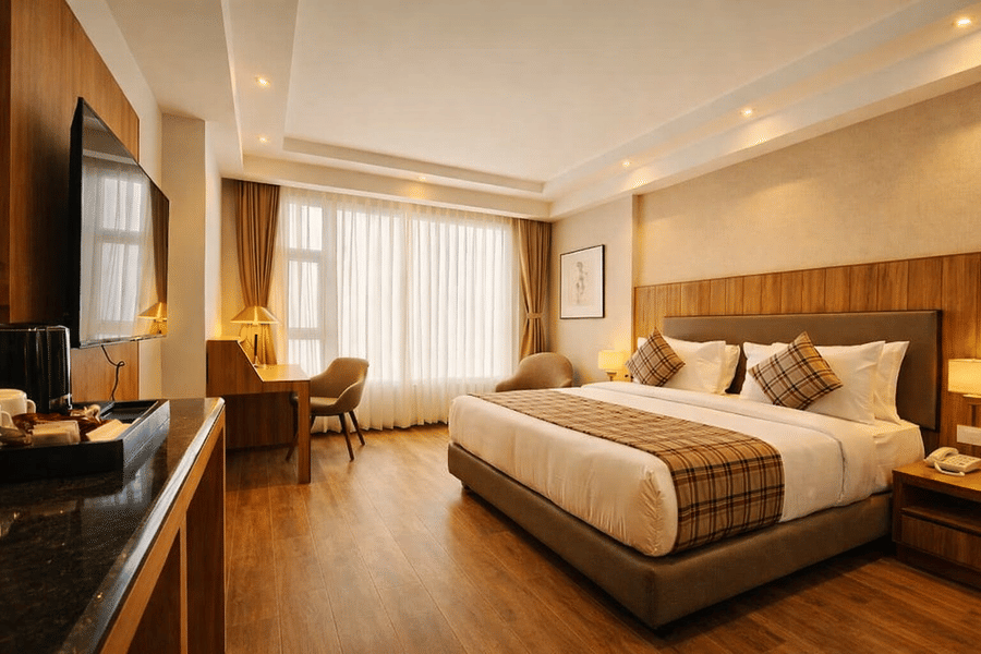 A bedroom at Pride Premier Darjeeling featuring a double size bed, a flat TV screen on the wall, a workstation with chair and desk, a sofa chair, and a large window with curtains drawn.