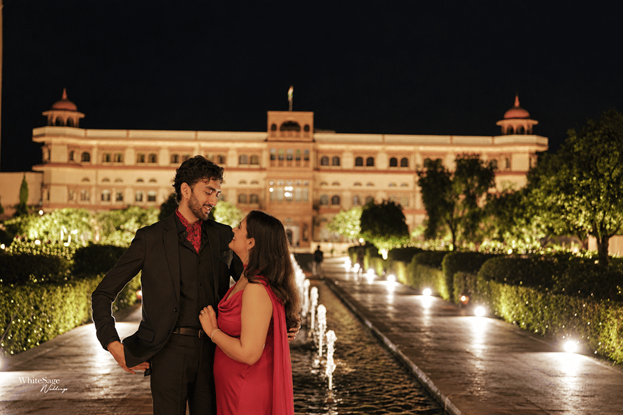 Couple posing outdoors beside a water fountain pathway in front of an illuminated facade at night at Umaid Palace.