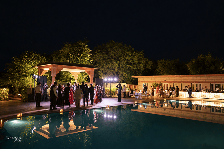 Night view of guests gathering around a swimming pool deck decorated with warm event lighting at Umaid Palace.