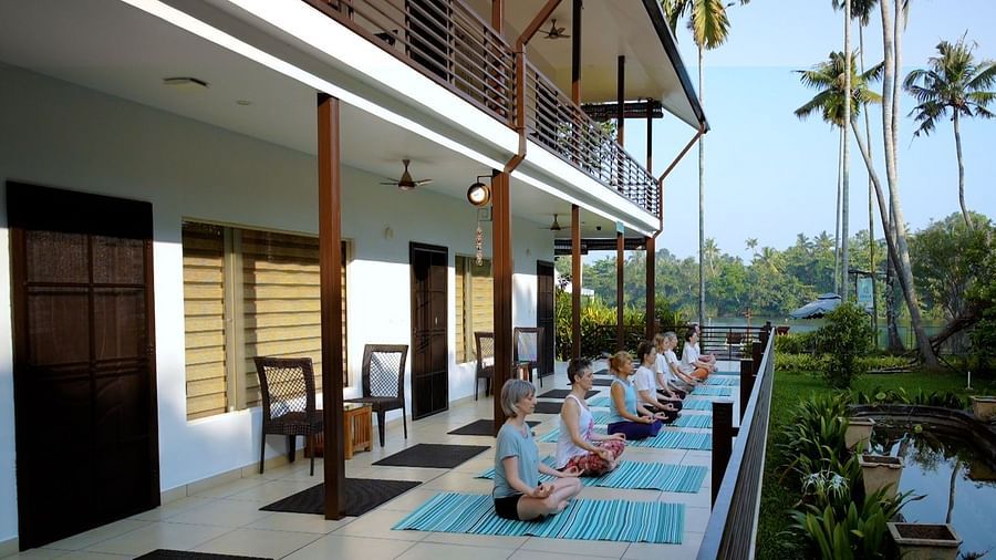A group of people sit in a row on yoga mats, practicing meditation or yoga on a long patio with a building and tropical foliage in the background.
