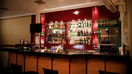 A view of a long wooden bar counter with upholstered chairs, a dark background with liquor bottles, and warm pendant lights at Hotel Horizon, Trivandrum.
