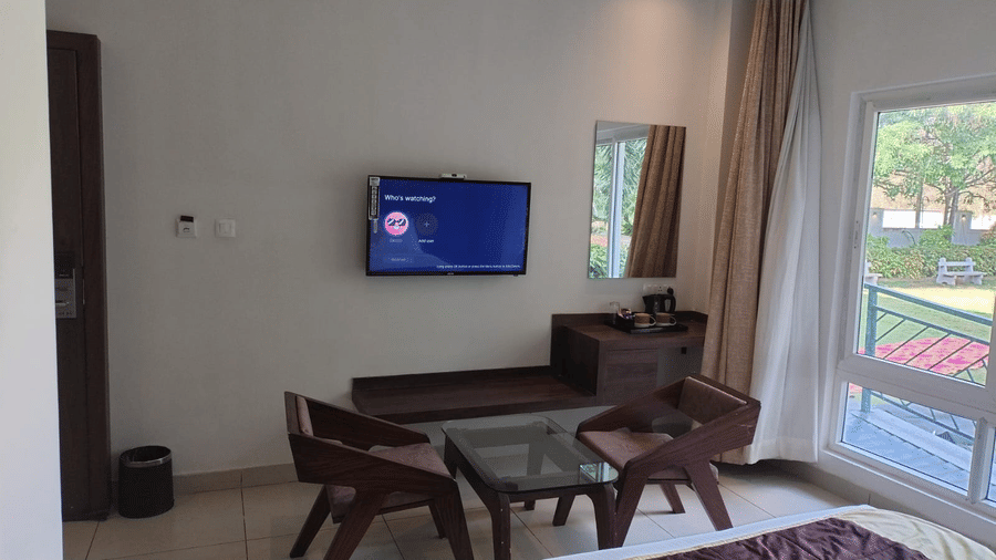 Standard Room at Cholaa Dynasty, Kumbakonam featuring a seating area with a wall-mounted TV, table with chairs, and balcony overlooking the lawn.