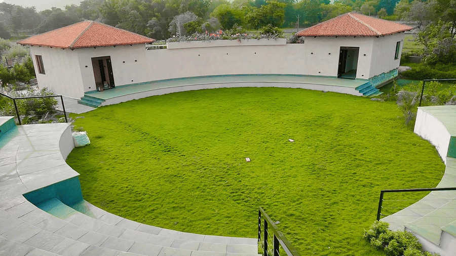 Large circular Golpo Bari green lawn with amphitheatre-style seating, surrounded by landscaped gardens and resort buildings, ideal for weddings, social gatherings, and outdoor events in Hotel Sonar Bangla Ajodhya Hills. 