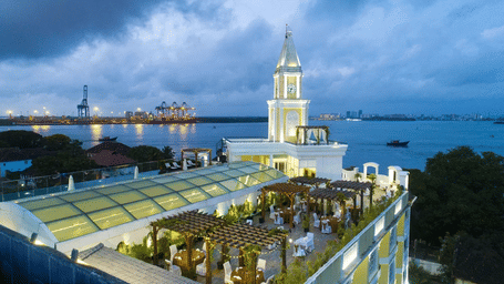 Aerial view of a rooftop dining terrace with pergolas and a clock tower, overlooking a harbor and city skyline at dusk.