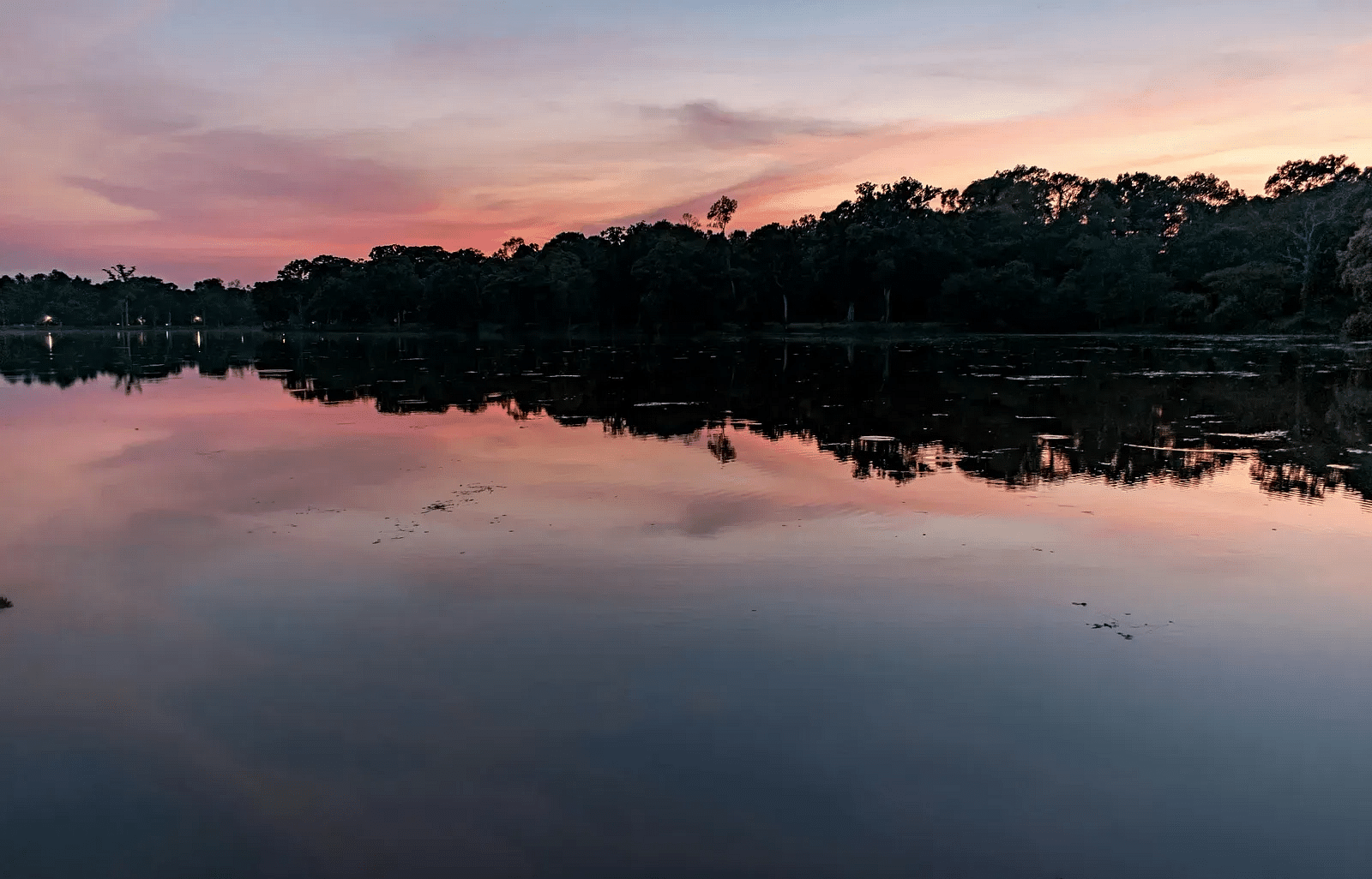 A serene lake at sunset with a colourful sky blending pink, purple, and orange hues, reflecting off the calm water surface, and a silhouette of trees lining the horizon.