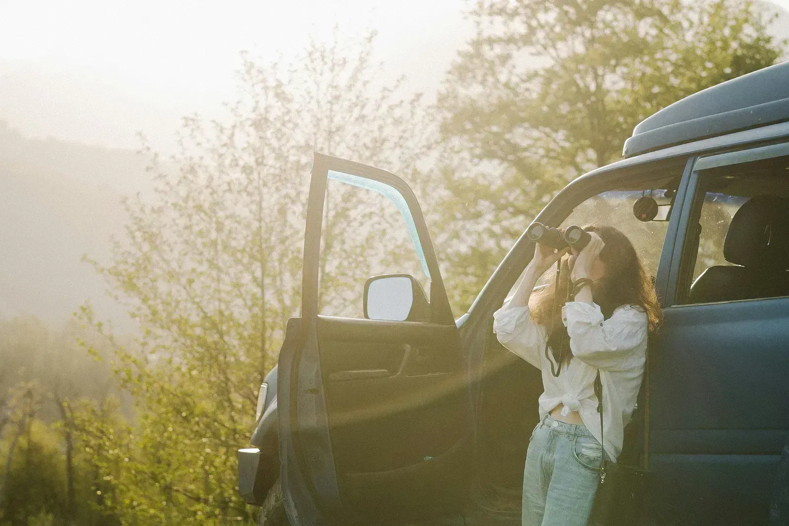 A lady looking into binoculars, leaning on a car surrounded by lush greenery.