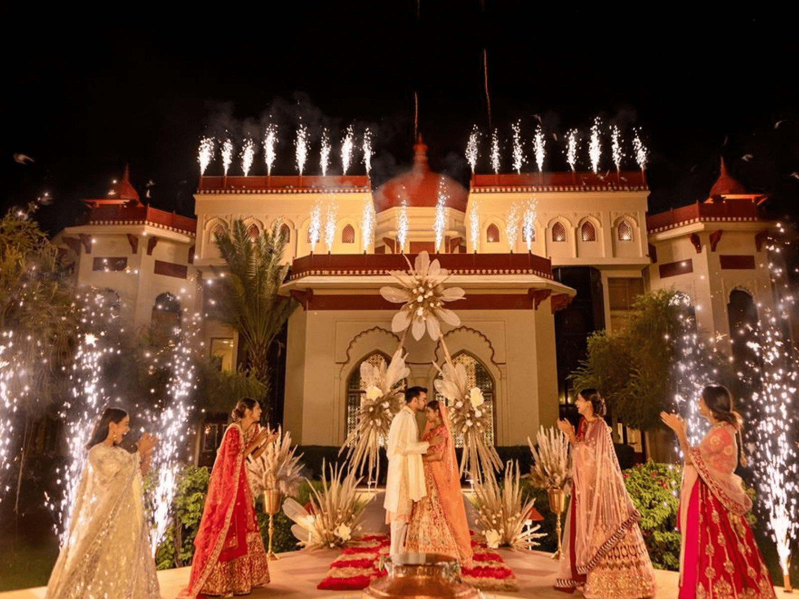 A couple standing under a floral archway at The Ummed Jodhpur, surrounded by friends and family holding sparklers, with the palace illuminated in the background.