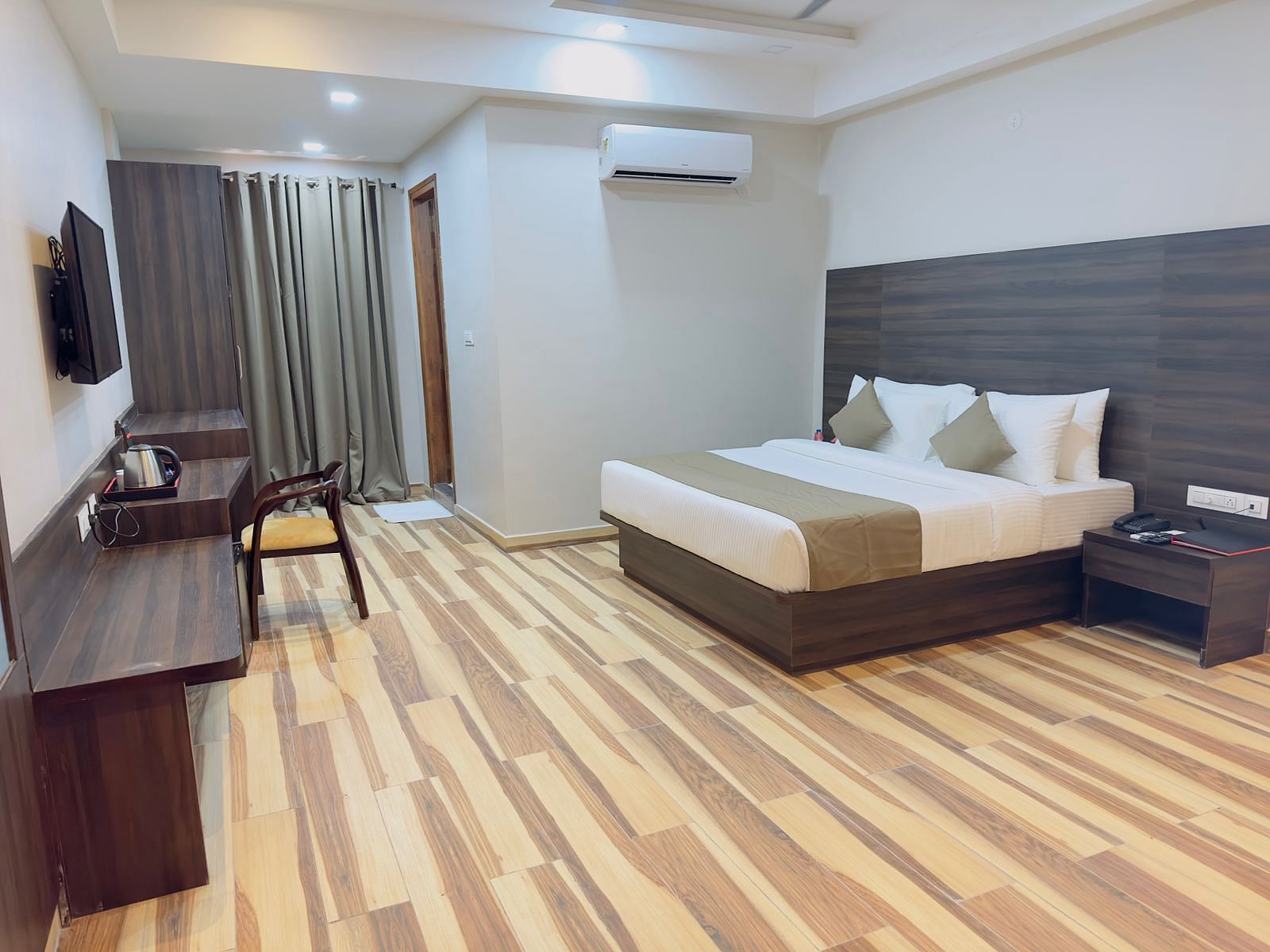 A room at Wild Wings Premium features a double bed with beige and white bedding, dark furniture, and wooden flooring - Kankidham Lords Inn