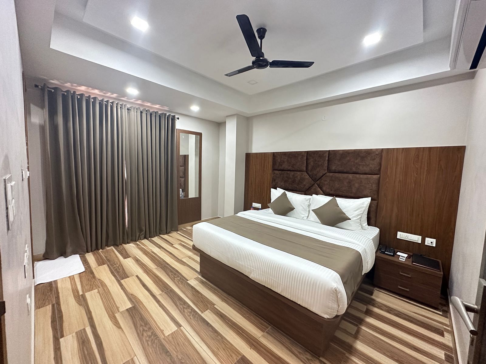 A room displays a double bed with white bedding, dark wooden headboard, and wooden flooring - Kankidham Lords Inn