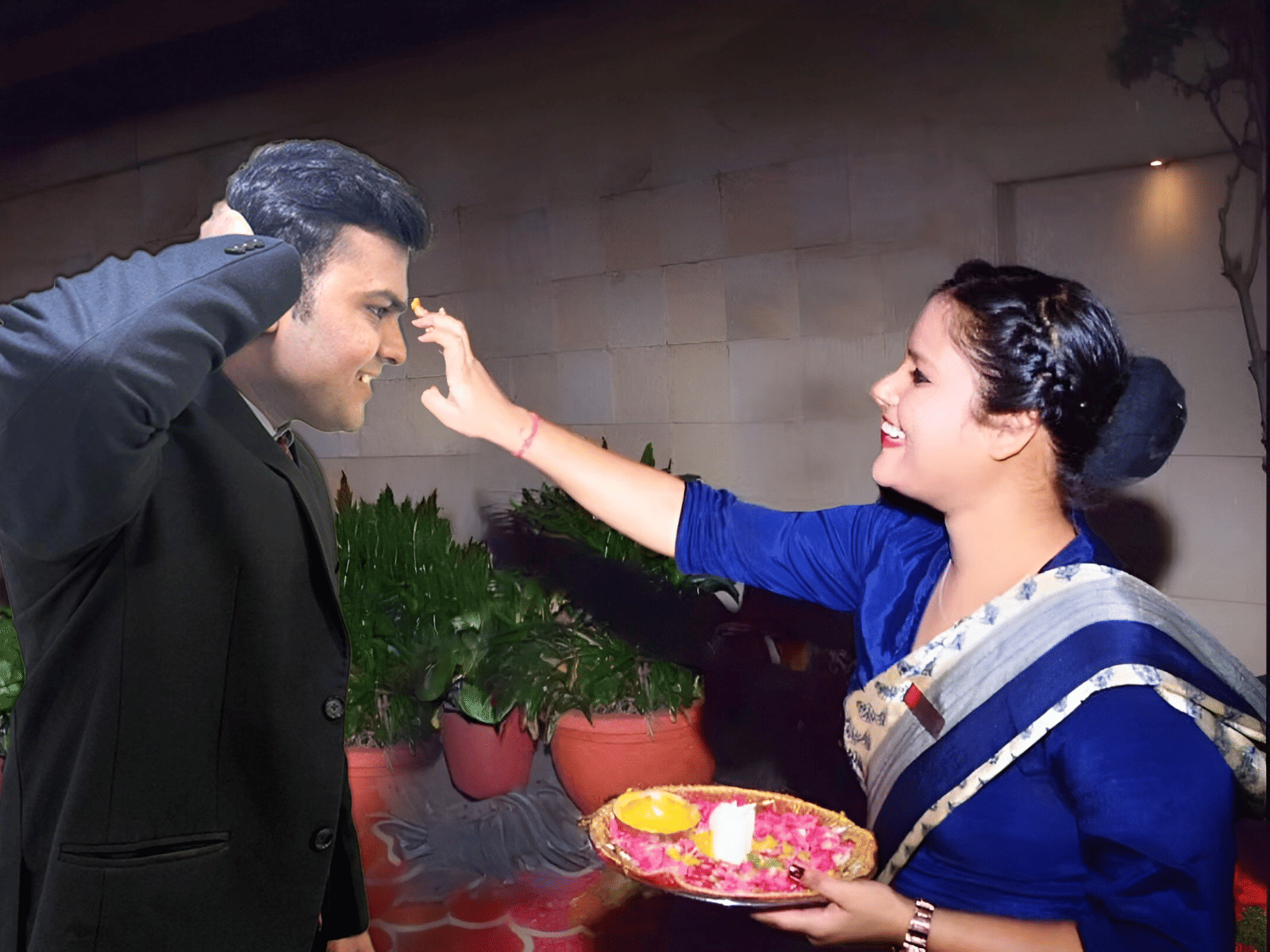 A woman in traditional attire applies a tilak on a man’s forehead during a festive or welcoming ceremony, both smiling warmly with a decorated plate of flowers and diya.