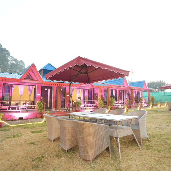 A set of chairs around a table with an umbrella to cover all who are seated, in front of some cottages