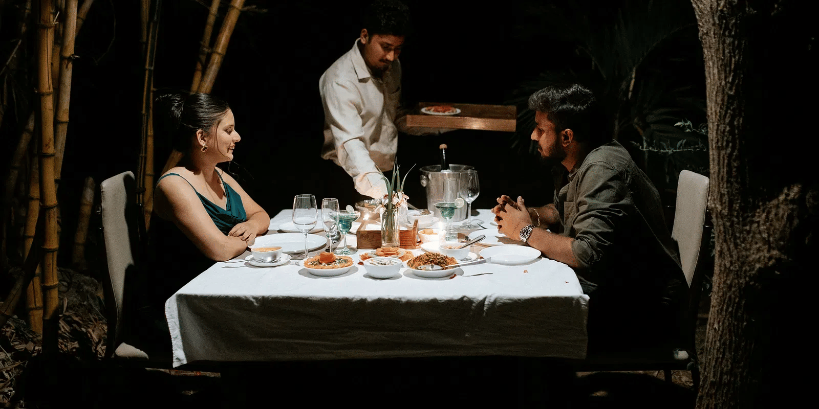 A couple having candle light dinner and a waiter of the resort serving them food in a romantic dim setting| Daksh Eden Greenz