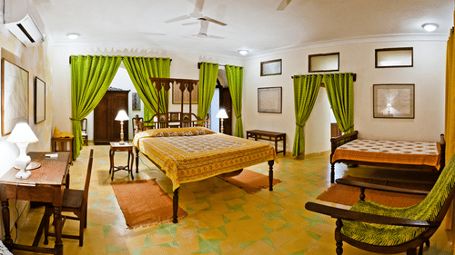 oom at Tijara Fort-Palace - 19th Century, Alwar contains two beds with patterned covers, bedside tables with lamps, a desk with chairs, seating, multiple windows with curtains, mirrors, and artwork.