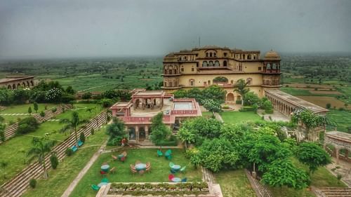 An aerial view shows Tijara Fort-Palace - 19th Century, Alwar nestled on a green hillside under a clear sky.
