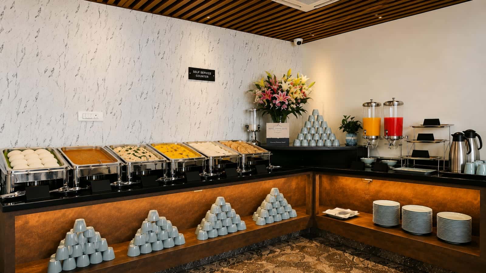 Self-service breakfast counter with chafing dishes, stacked plates and bowls, and disposable cups