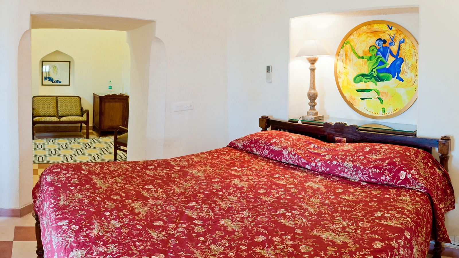 A guest room at Neemrana's Tijara Fort Palace features a bed with a patterned cover, a bedside table with a lamp and artwork, and an arched doorway leading to another area with seating and a mirror.