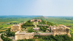 An aerial view shows Tijara Fort-Palace - 19th Century, Alwar nestled on a green hillside under a clear sky.Facade_ Facade_Tijara Fort Palace_Hotel In Rajasthan_ Palace Hotel In Rajasthan 111