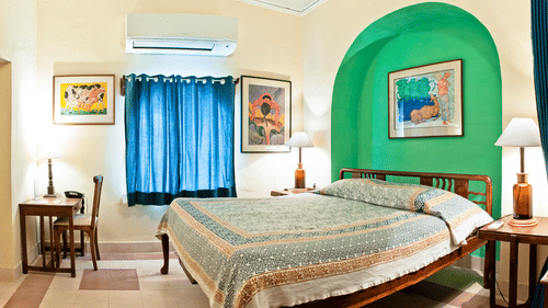 A guest room at Tijara Fort-Palace - 19th Century, Alwar contains a bed with patterned linens, bedside tables with lamps, artwork on the walls including one in a green arched alcove, a window with blue curtains, a desk with a chair, and an air conditioning