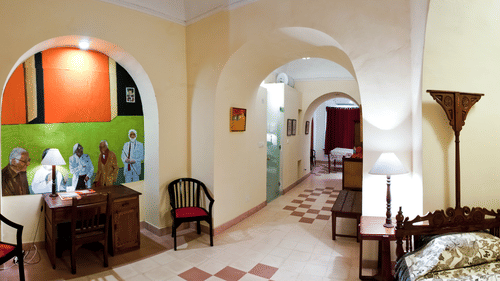 A hallway at Tijara Fort-Palace - 19th Century, Alwar features arched openings, tiled flooring, a floor lamp, a table lamp, chairs, a desk with a mural in an alcove, and glimpses into guest rooms.