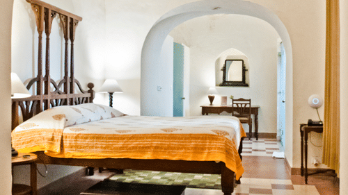 A guest room at Tijara Fort-Palace - 19th Century, Alwar includes a bed with patterned and solid-toned linens, bedside tables with lamps, a mirror, a desk with a chair, and arched doorways.