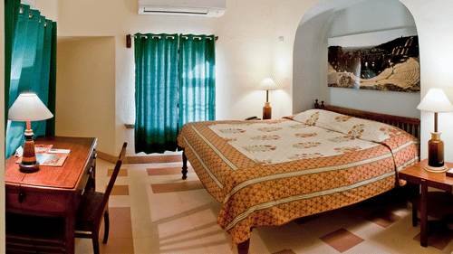 A guest room at Tijara Fort-Palace - 19th Century, Alwar features a bed with patterned linens, bedside tables with lamps, a desk with a chair, windows with curtains, artwork in an arched alcove, and an air conditioning unit.
