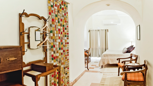 A hallway at Tijara Fort-Palace - 19th Century, Alwar features a dressing table with a mirror and coat hooks, a patterned curtain, arched doorways, and a glimpse into a bedroom with a bed and seating.