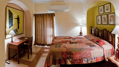 A guest room at Neemrana's - Tijara Fort Palace features a bed with a patchwork cover, bedside tables with lamps, a desk with a lamp and chair, artwork, a window with curtains, and air conditioning.