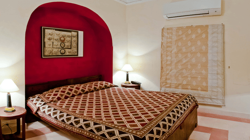 A guest room at Neemrana's Tijara Fort Palace features a bed with a patterned cover, bedside tables with lamps, artwork in a red arched alcove, a decorative textile hanging, and air conditioning.