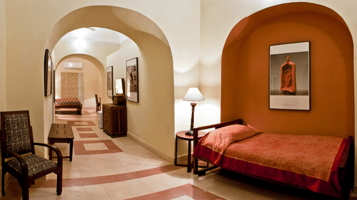 A hallway at Tijara Fort-Palace - 19th Century, Alwar features arched openings, tiled flooring, a chair, a bench, framed artwork, and a single bed with a bedside table and lamp in an orange arched alcove and a glimpse into another guest room is visible.