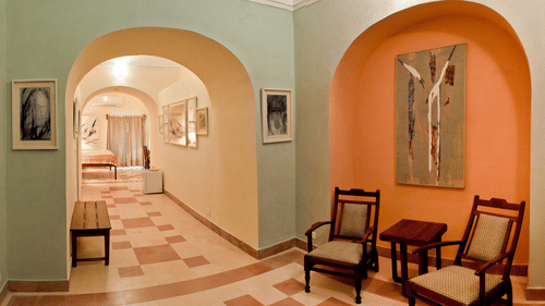 A hallway at Tijara Fort-Palace - 19th Century, Alwar features arched openings, tiled flooring, a bench, framed artwork on the walls, and a seating area with chairs and a small table in an orange arched alcove. A glimpse into a guest room is visible.