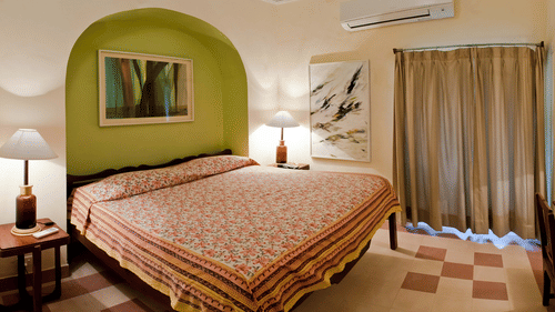 A guest room at Tijara Fort-Palace - 19th Century, Alwar includes a bed with patterned linens, bedside tables with lamps, artwork in a green arched alcove, another piece of artwork, a window with curtains, and air conditioning.