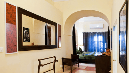 A hallway at Tijara Fort-Palace - 19th Century, Alwar features a large mirror, artwork, a towel rack, seating, tiled flooring, and an arched doorway leading to a guest room with a bed, curtains, and air conditioning.