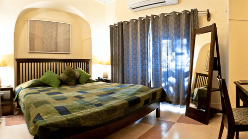 A guest room at Tijara Fort-Palace - 19th Century, Alwar includes a bed with a patchwork cover and green pillows, bedside tables, a full-length mirror, a desk with a chair, a window with striped curtains, an air conditioning unit, and tiled flooring.
