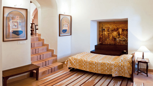 A guest room at Tijara Fort-Palace - 19th Century, Alwar features a bed with a patterned cover, a bedside table with a lamp, a built-in seating area with artwork above, a bench, tiled stairs, and framed artwork on the walls.