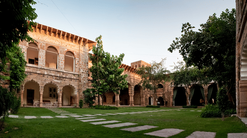 The courtyard at Tijara Fort-Palace - 19th Century, Alwar shows stone buildings with arched openings surrounding a green lawn with trees and stone pathways.