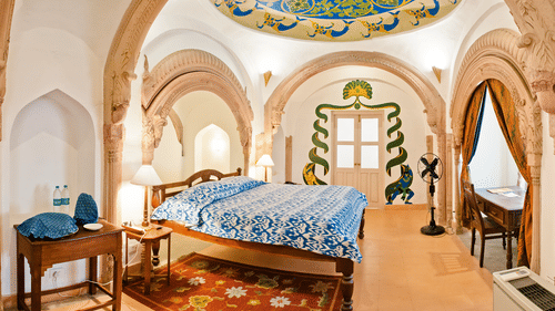 Suite at Tijara Fort-Palace - 19th Century, Alwar showcases a bed with blue and white patterned linens, bedside tables with lamps, a rug, arched doorways, a decorated ceiling, a window with curtains, a desk with chairs, and unique wall decorations.