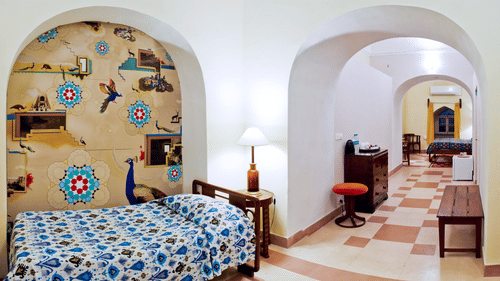 A guest room at Tijara Fort-Palace - 19th Century, Alwar includes a bed with patterned linens, a bedside table with a lamp, a decorated arched alcove, and an arched opening leading to a tiled hallway with furniture.